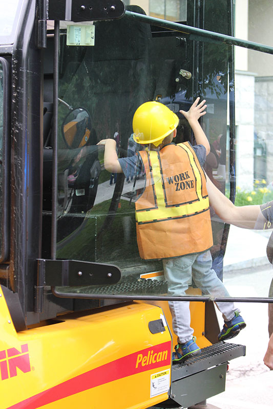 Child wearing yellow hard hat and orange "work zone" vest climbing up on a construction truck.