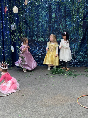 Three young children dressed as princesses stand in background as one young child dressed as a princess kneels in front of them.