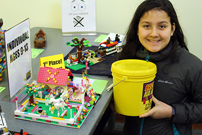 Child holding a yellow brick bucket in front of a Lego construction.