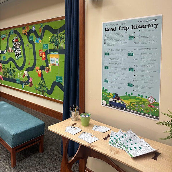 Side view of Cary Library's Idea Wall with the road map in background and a "Road Trip Itinerary" poster on the wall above a table with sheets and writing utensils for patrons to participate.